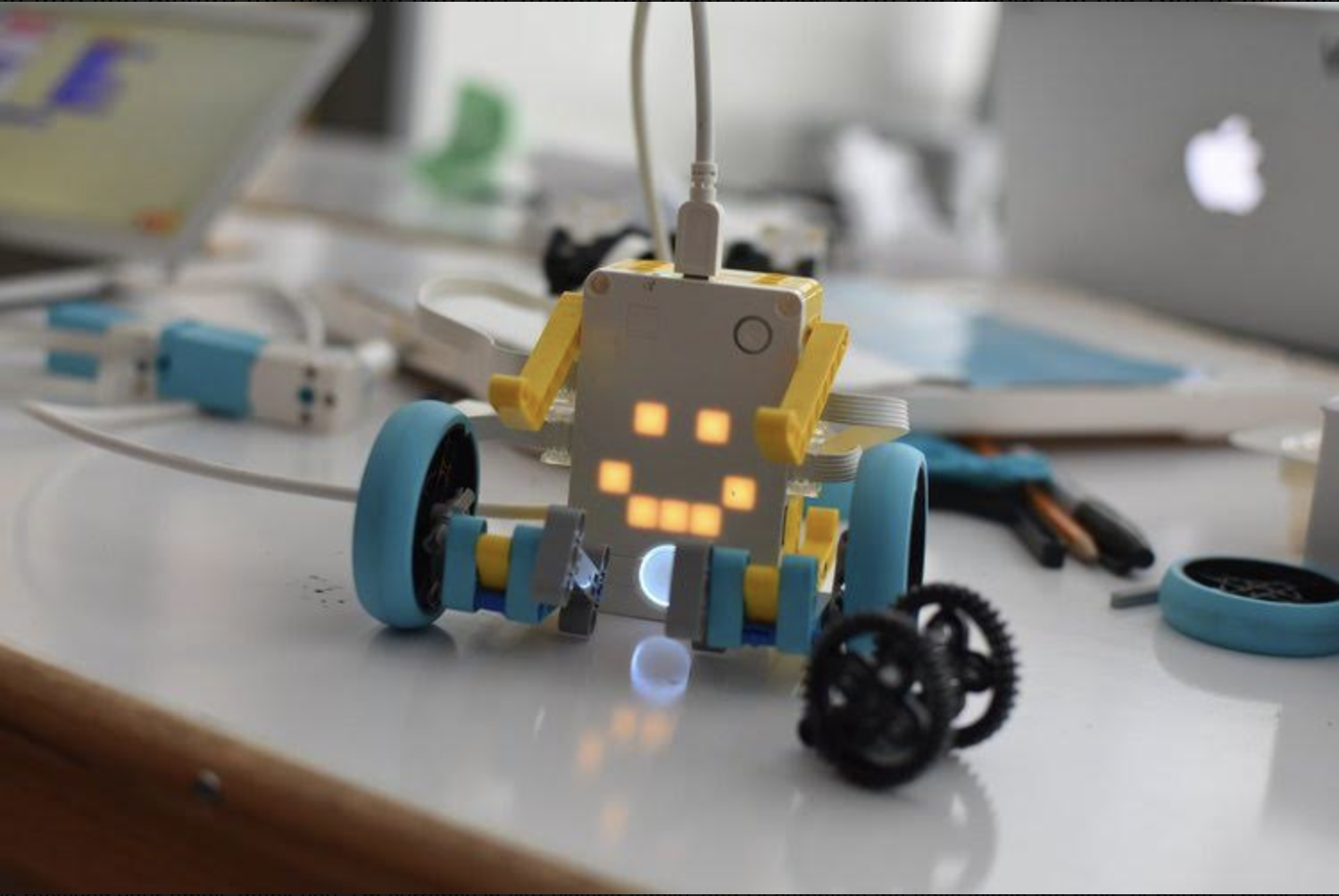 A close-up view of a small, cheerful-looking robot with glowing eyes on a desk scattered with various robotics parts and tools. In the background, there's a blurred view of a laptop with the Apple logo, indicative of the technology-rich learning environment at Court Street Arts.